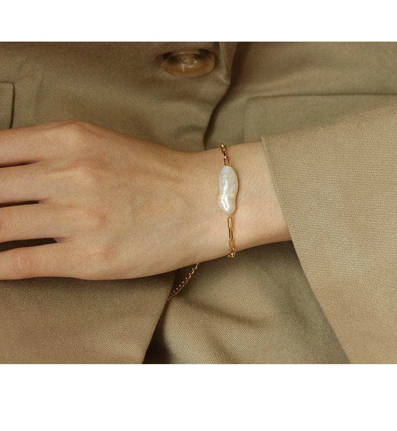 Gold bracelet with a large pearl worn on a wrist against a beige background