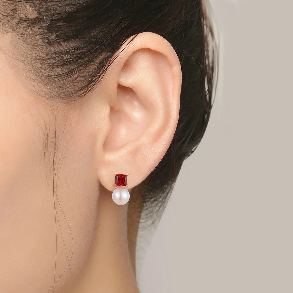 Close-up of an ear wearing a red gemstone and pearl earring against a neutral background