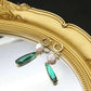 Gold earrings with green teardrop and pearl charms in front of a gold mirror.