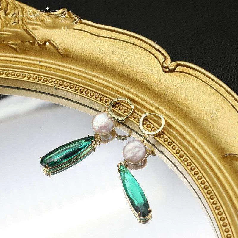 Gold earrings with green teardrop and pearl charms in front of a gold mirror.
